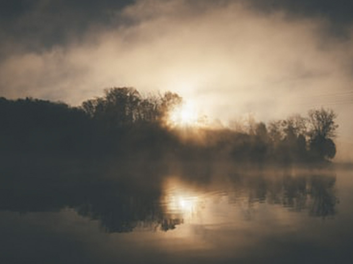 A foggy lake with trees and sun shining through the clouds.