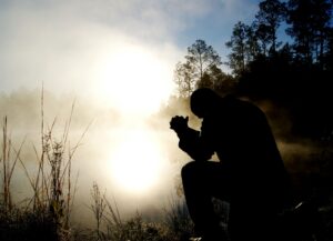 A man kneeling down in the grass with his hands clasped.